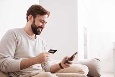 portrait of a cheerful man shopping online with credit card and mobile phone while sitting on a couch at home
