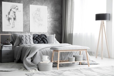 charcoal sketches hanging on a concrete wall near the window in bright bedroom interior with wooden lamp and bench in front of the bed