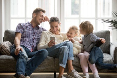 cheerful young family with kids laughing watching funny video on smartphone sitting on couch together, parents with children enjoying playing games or entertaining using mobile apps on phone at home