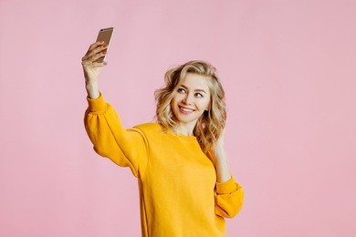 close-up portrait of cheerful  young caucasian female with curly blonde hair, in yellow sweater poses on a pink background. woman takes a selfie on the camera of the smartphone