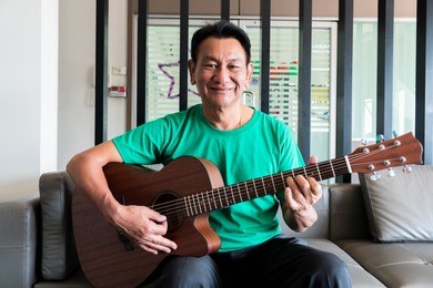 portrait of old man with acoustic guitar with happiness retirement, on sofa in room, concept music therapy.