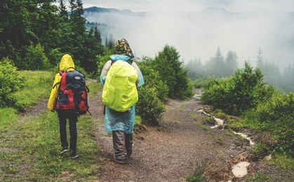 two hikers tourist in raincoat walking on trail to green mountain forest in the fog with the yellow backpack in rainy weather.