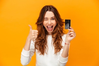 cheerful brunette woman in sweater showing thumb up and holding credit card while winks and looking at the camera over yellow background
