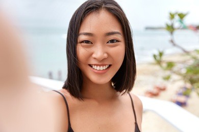 pretty young asian woman takes photo on camera og unrecognizable gadget, poses against sea or ocean horizon, takes picture during free time, enjoys good rest. outdoor shot of cute chinese girl