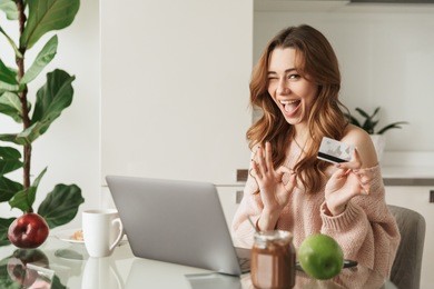 portrait of a delighted young woman holding credit card while sitting with laptop computer at the table at home