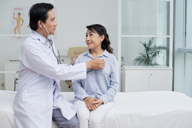 profile view of confident middle-aged therapist wearing white coat using stethoscope while examining senior patient, interior of modern ward on background