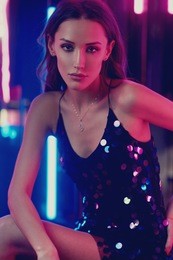 waist-up portrait of young gorgeous woman with long brunette hair wearing elegant black sequin strap cami dress. beautiful slender female model posing against glowing neon lights on background