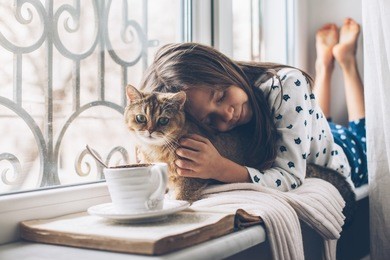 child in pajamas relaxing on a window sill with pet. lazy weekend with cat at home. cozy scene, hygge concept.