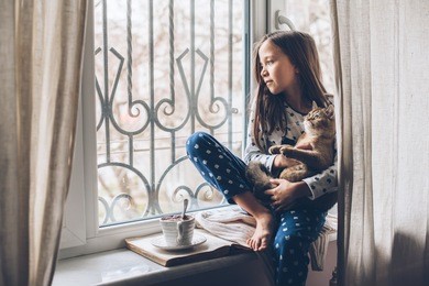 child in pajamas relaxing on a window sill with pet. lazy weekend with cat at home. cozy scene, hygge concept.