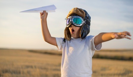 fun little boy wearing helmet and dreams of becoming an aviator while playing a paper plane at sunset