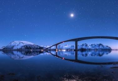 amazing bridge and starry sky with beautiful reflection in water. night landscape with bridge, snowy mountains, blue sky with moon and bright stars reflected in sea. winter in lofoten islands, norway 