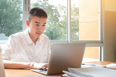 technology learning concept: asian young man student learning and check timetable for projected in laptop computer at busy table in high school with chart document files on wood desk near window.