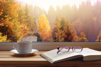 
a cup of fragrant coffee with an open book and glasses on a wooden windowsill against the background of the autumn sunny forest outside the window