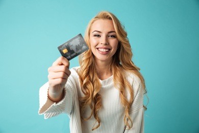 portrait of a smiling young blonde woman in sweater showing credit card isolated over blue background
