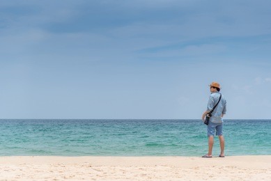 young asian man photographer standing on the beach. sea and sky horizon in background. travel holiday or vacation in summer season concept