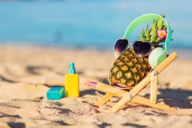 ripe attractive girl-pineapple in stylish headphones lying on sunbed on the sand against turquoise sea. listening music, relaxing. wearing sunglasses. tropical summer vacation concept. sunbathing