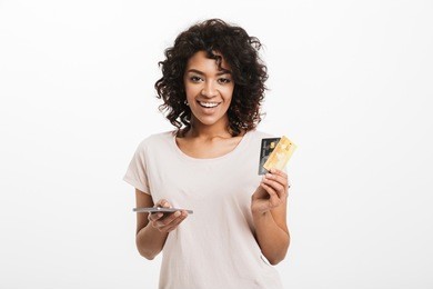 portrait of a smiling young afro american woman holding mobile phone and showing credit cards isolated over white background