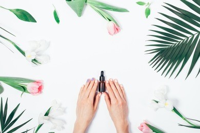 top view of woman holding cosmetics bottle with pipette. spring white and pink flowers, summer palm branch. flat lay composition on white background for beauty blog.