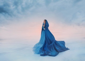 the snow queen in a blue raincoat that flutters in the wind. a traveler on a background of sunrise or sunset, and a frozen valley covered with snow and clouds, a place where heaven meets the earth.