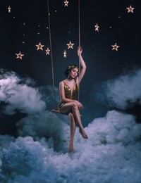 an asterisk girl, swinging on a rocker in the clouds, among the stars and the night sky. the greek goddess in a golden dress and in a laurel crown looks at the world from heaven. art's photo