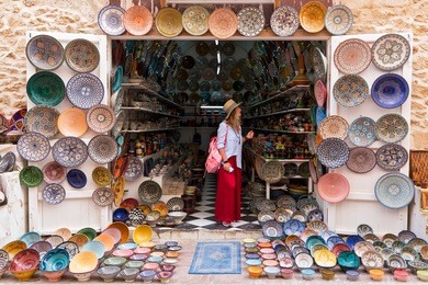 young blonde woman in a long red skirt, hat and with pink backpack is standing in a small shop and looking at moroccan colorful plates.