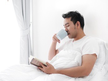 happy asian man reading a book and drinking coffee on bed, man lifestyle on weekend concept.