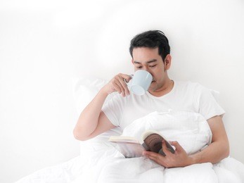 happy asian man reading a book and drinking coffee on bed, man lifestyle on weekend concept.