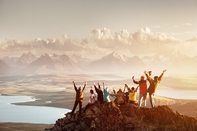 big group of people having fun in success pose with raised arms on mountain top against sunset lakes and mountains. travel, adventure or expedition concept