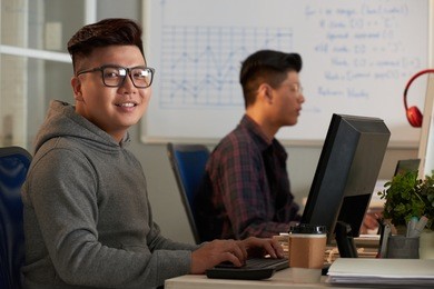 smiling asian student wearing eyeglasses looking at camera while sitting in front of modern computer during ict class, portrait shot