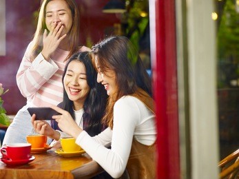 three happy beautiful young asian women sitting at table chatting talking laughing playing with cellphone in coffee shop or tea house, shot through window glass.