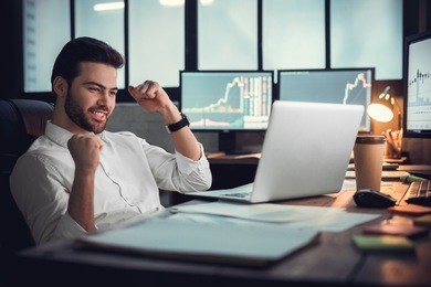 young male trader at office work concept sitting looking at laptop happy