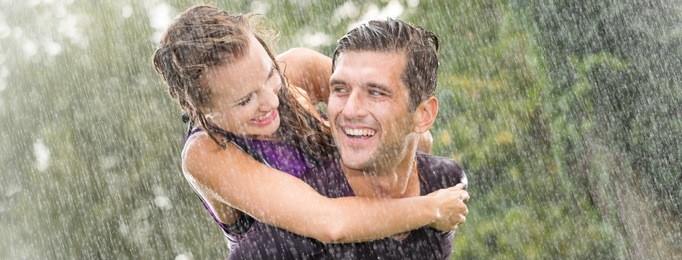 smiling young man holding pretty girlfriend on his back during heavy summer rain