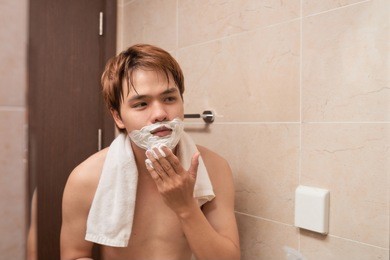 handsome young asian man is shaving his face and looking at the mirror.