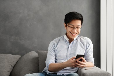 portrait of a happy young asian man using mobile phone while sitting on a couch at home