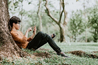 happy casual asian man using a smart phone outdoor sitting under tree in park