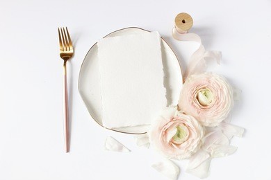 feminine wedding, birthday desktop mock-up scene. porcelain plate, blank craft paper card, silk ribbon, blush pink persian buttercup flowers and fork. white table background. flat lay, top view.