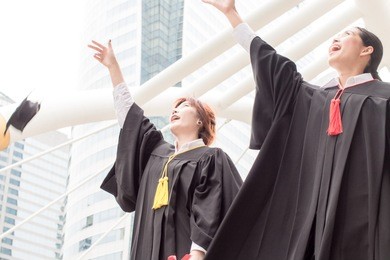 best friend with young female students dressed in black graduation gown or graduates with congratulations with graduation hats is standing, smiling and holding diplomas with business city background.