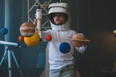cute little boy wearing cardboard astronaut helmet flying toy rocket through planets, cardboard spaceship rocket in the background