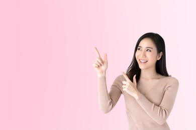 beautiful young casual asian woman smiling and pointing hands to empty space aside, studio shot on pink background
