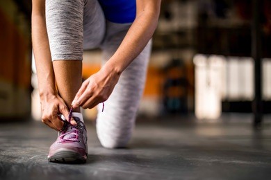 woman tying her shoe in an open empty gym, preparing for workout or run, wearing gray leggings and royal blue top.