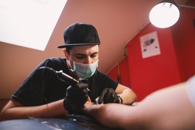 young focused tattoo artist is inking customers arm carefully in his shop.