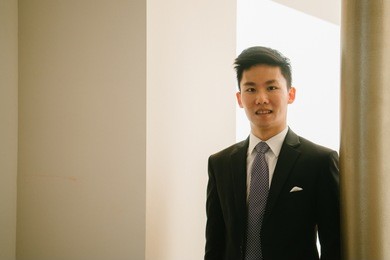 portrait of a young, chinese lawyer in a suit standing in a hallway and smiling.