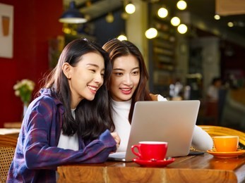 two happy young asian women sitting in coffee shop looking at laptop computer together.