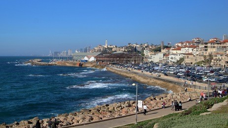 view from hill at jaffa portand buildings, mediterranean sea and tel aviv skyscrapers in background, waves on water, shore with stones, car park with cars, people walk along promenade, sunny, blue sky