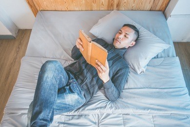 a young asian man reading a book on a bed