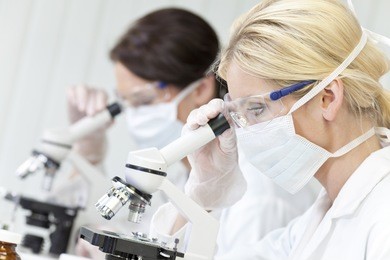 a blond female medical or scientific researcher or doctor using her microscope in a laboratory with her colleague out of focus behind her.