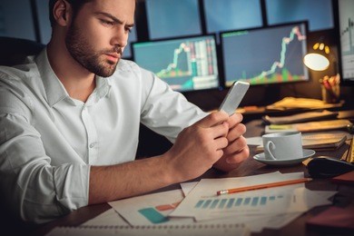 serious trader using mobile phone app on smartphone for checking stock trading data analysis concept, focused businessman holding cellphone working in office with financial graph on computer monitors