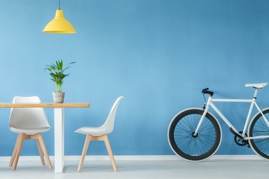 minimal, modern interior with two chairs, a bicycle, a table with a plant on it and a yellow lamp above, against blue wall