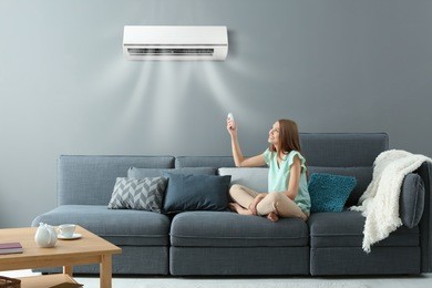 young woman switching on air conditioner while sitting on sofa at home