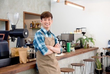 cheerful young asian man entrepreneur standing with arm crossed at counter in his own coffee shop